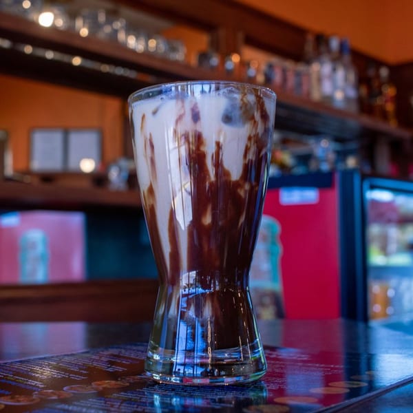 A tall glass of iced coffee with chocolate syrup swirls sits on a dark, reflective bar.