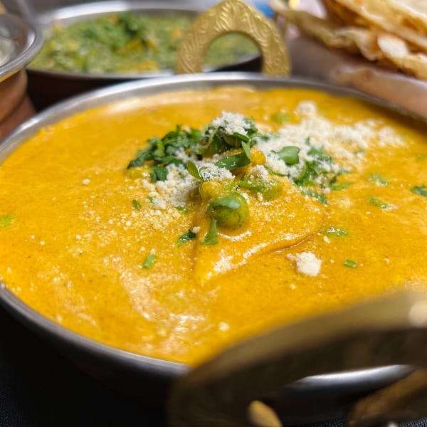 Close-up of a vibrant orange Indian curry, garnished with fresh herbs and grated cheese in a metal bowl.