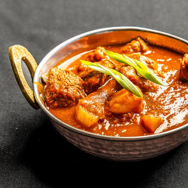 A metal bowl of Indian curry with meat and potatoes, garnished with green onions.
