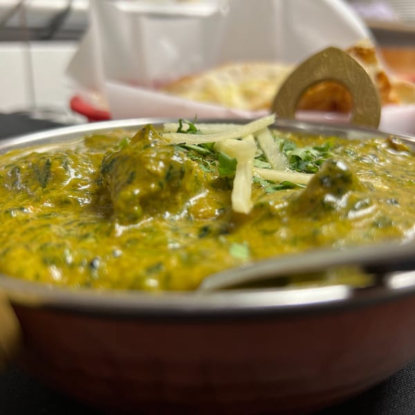 Close-up of vibrant green Indian curry, garnished with fresh herbs and ginger, with naan bread in background.