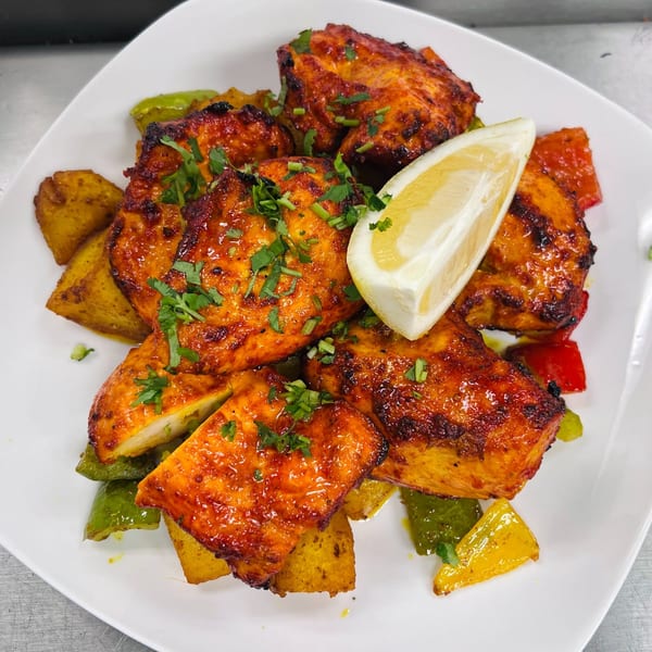 Tandoori chicken tikka with bell peppers, potatoes, cilantro, and a lemon wedge on a white plate.
