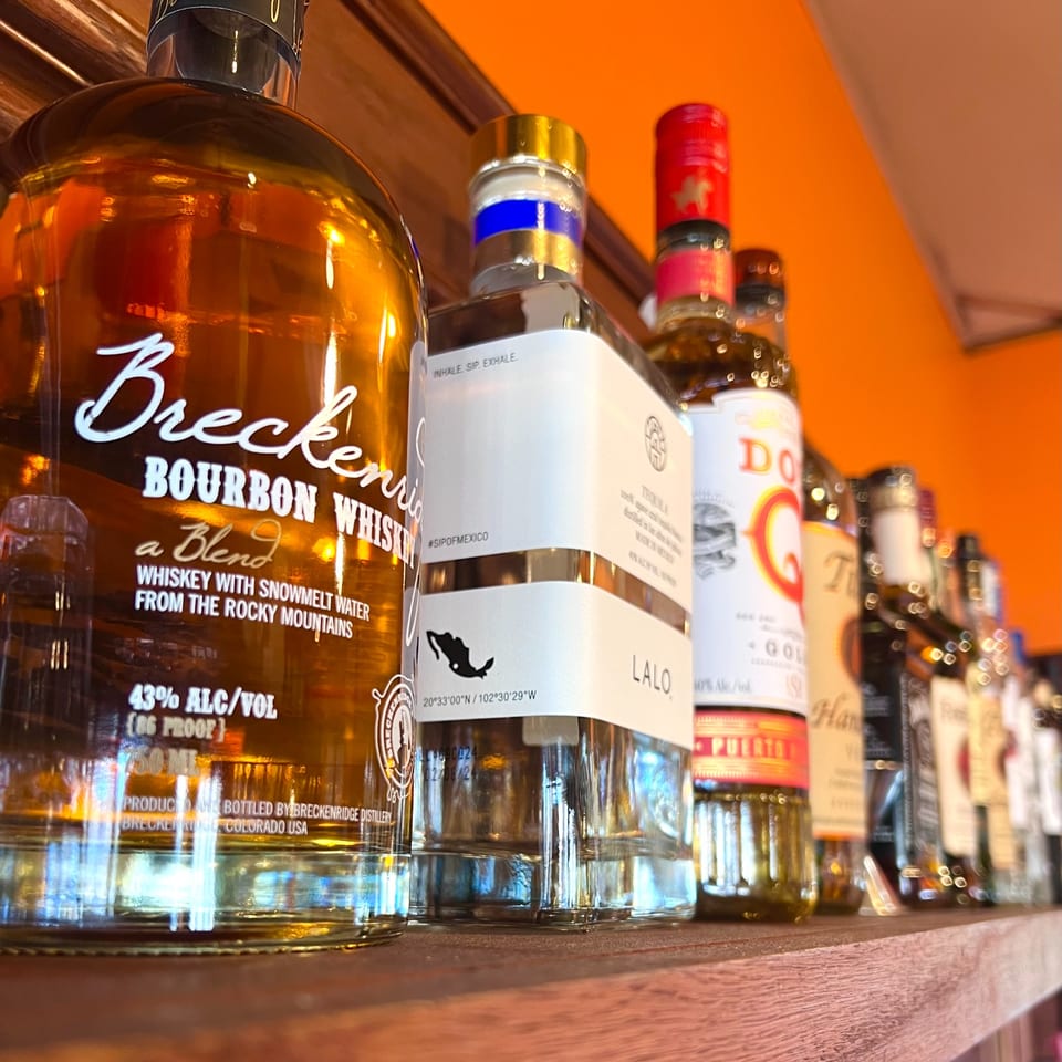 A close-up of various liquor bottles, including Breckenridge Bourbon, on a wooden shelf against an orange wall.