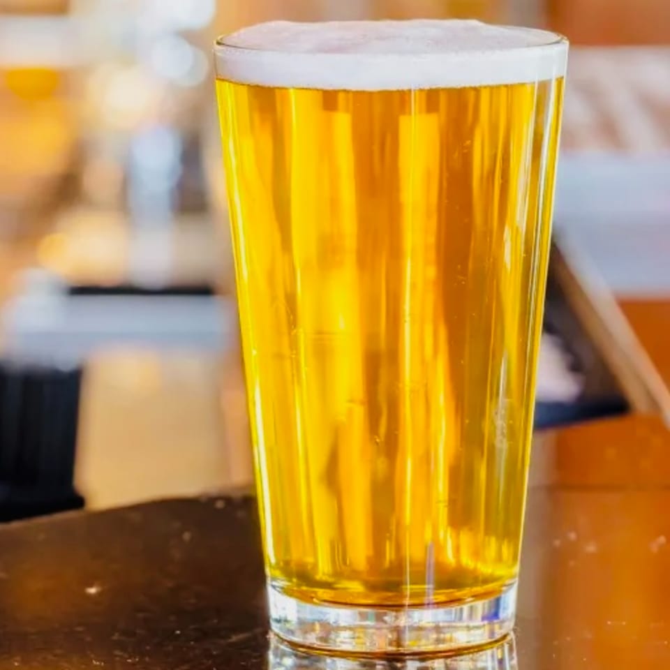 A pint glass of golden beer with a foamy head sits on a dark bar top.