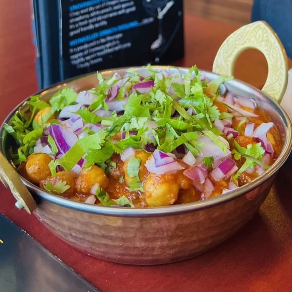 A close-up of a metal bowl filled with Indian chana masala, topped with chopped red onions and cilantro.