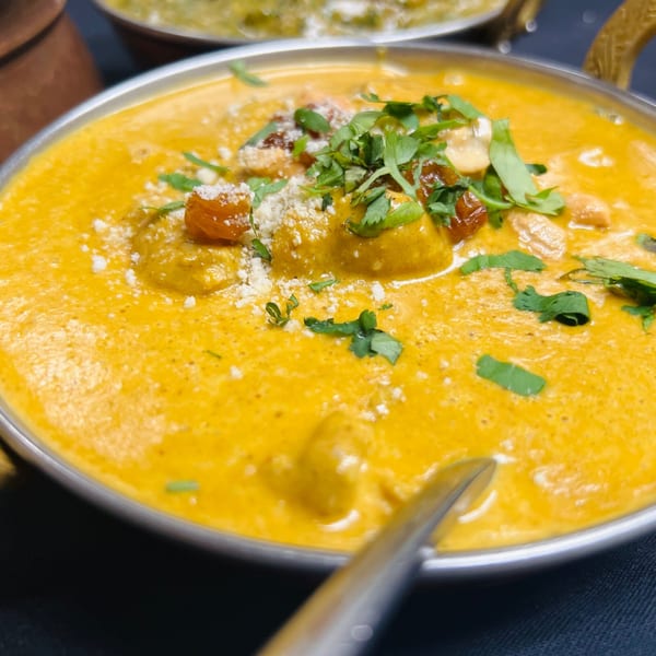 Close-up of creamy yellow Indian curry with cilantro, nuts, and dried fruit in a metal bowl.