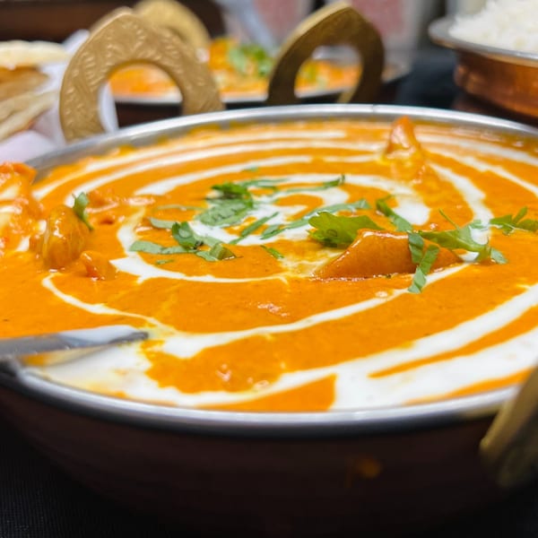 Close-up of a rich orange Indian curry with white cream swirls and green herb garnish in a metal bowl.