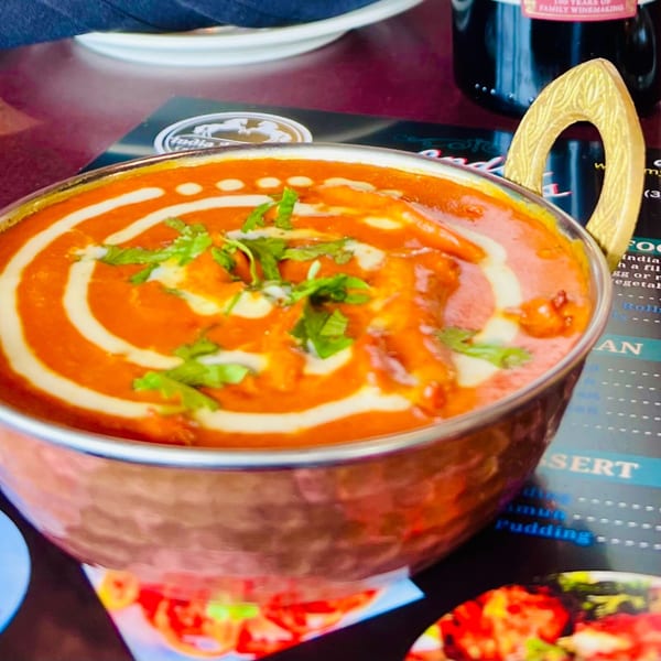 A copper bowl of vibrant orange Indian curry, garnished with cream swirls and fresh cilantro, on a restaurant table.