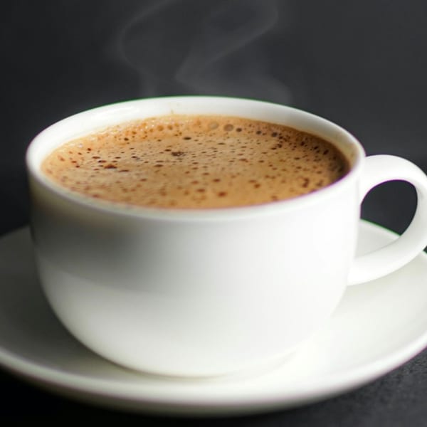 Steaming hot chai tea with frothy top in a white mug and saucer against a dark background.