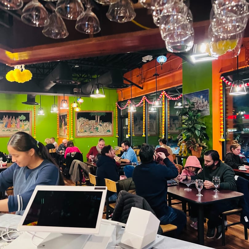 Diners enjoying a vibrant restaurant interior with green walls, garlands, and hanging wine glasses.