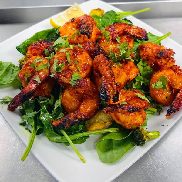 Tandoori shrimp with charred edges, garnished with cilantro and lemon, served on a bed of spinach.