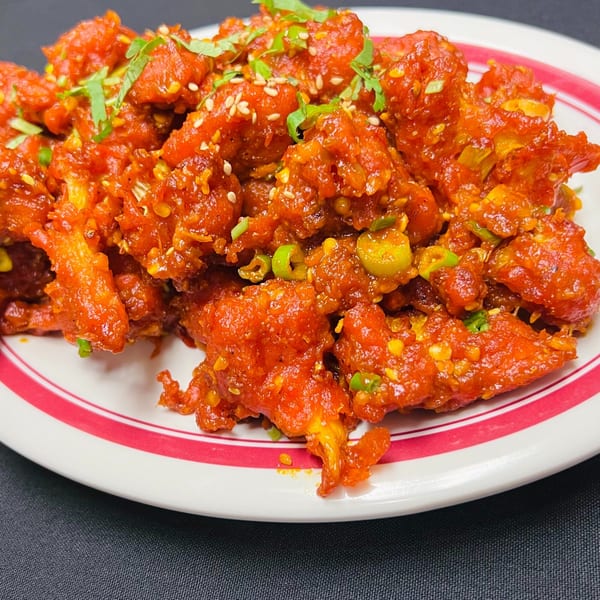Bright red chili chicken, garnished with green herbs and sesame seeds, on a white plate with red stripes.